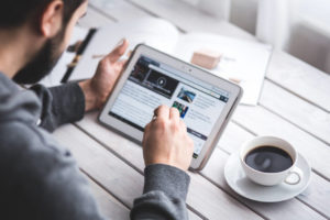 Blick über die Schulter eines Mannes, der ein Tablet bedient. Neben dem Tablet steht ein Kaffee auf dem Tisch.