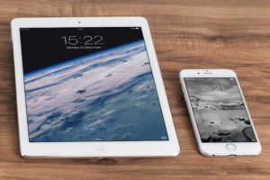 Ein iPad und ein iPhone liegen auf einer Holzplatte. Auf den Geräten sind unterschiedliche Startbildschirme erkennbar.